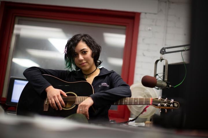 Emily Spector has taken songwriting classes on both sides of the river. She is sitting here holding a guitar