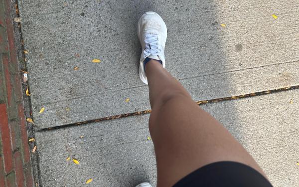 A girl's tan leg wearing black running shorts and white running sneakers, mid stride on a concrete sidewalk.